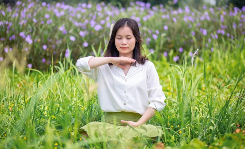 Žena sedi na travnatoj livadi u pozi meditacije sa rukama na kolenima, okružena prirodom