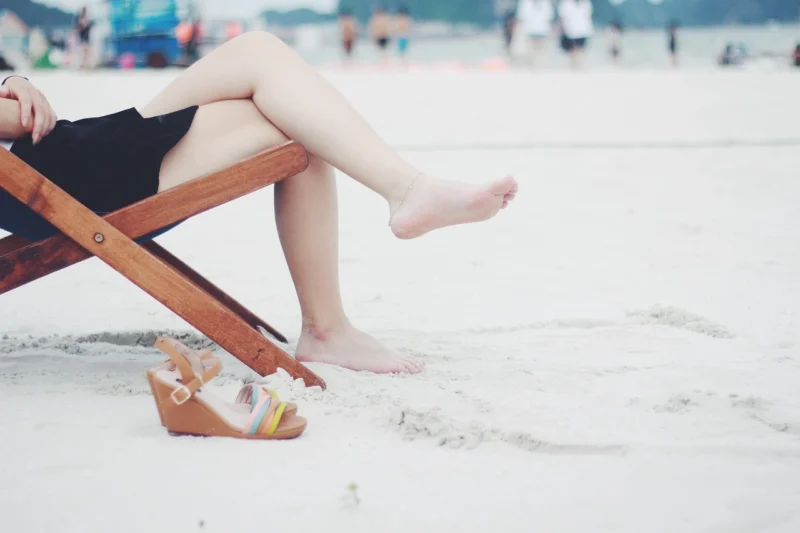 Žena sedi na drvenoj stolici na peščanoj plaži, noge joj dodiruju pesak dok gleda u more